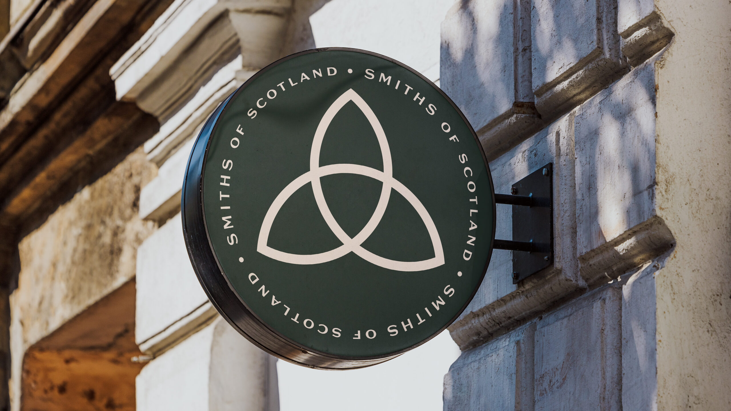 Round hanging sign on a stone wall: the redrawn Celtic knot inside a Smiths of Scotland roundel, cream on green